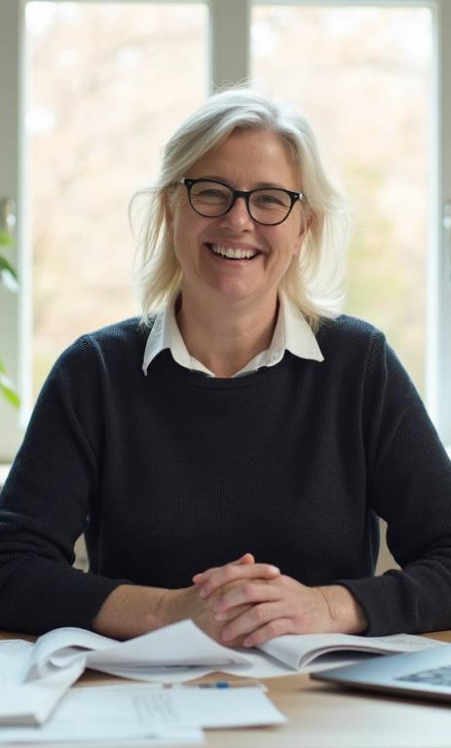 Donna professionista sorridente in ufficio, seduta alla scrivania con laptop e documenti, ambiente moderno con piante da interno.